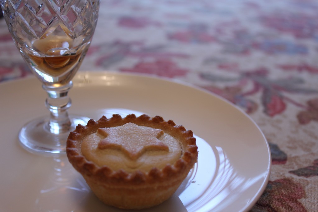 A mince pie and glass of sherry sitting on a white ceramic plate, on a cream and red-flower patterned tablecloth.